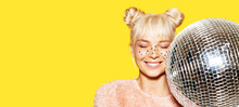 Close-up Portrait Of Happily Smiling Girl With Two Lovely Buns Holding Disco Ball. Cheerful Lady In Pink Sweater. Festive Makeup And Party Concept. Isolated On Yellow Background