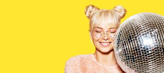 Close-up portrait of happily smiling girl with two lovely buns holding disco ball. Cheerful lady in pink sweater. Festive makeup and party concept. Isolated on yellow background