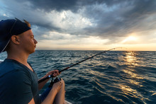 Young Man Fishing By Spinning In Boat In Opened Sea. Beautiful Sunset Seascape.
