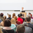 © kasto - Business and entrepreneurship symposium. Female speaker giving a talk at business meeting. Audience in conference hall. Rear view of unrecognized participant in audience. Copy space on whitescreen.