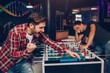 © estradaanton - Youn bearded happy man cheering at soccer table in playing room. He wins. Another guy stand behind. He looks upset.