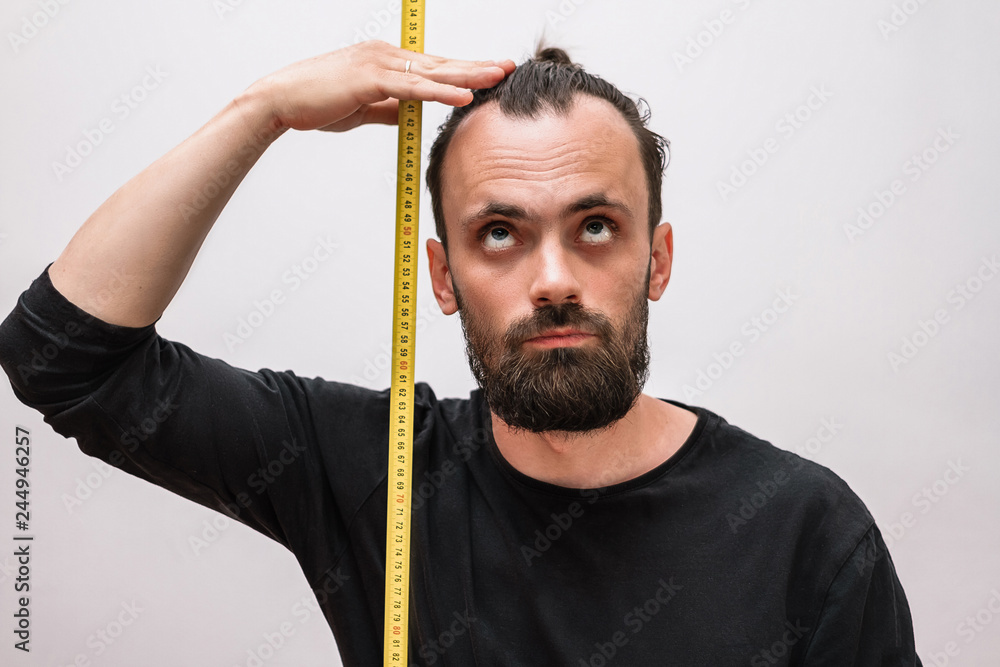 a man with a beard and a black t-shirt makes measurements of his height with a tape measure - obrazy, fototapety, plakaty 