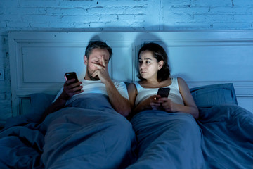 Sad and bored couple addicted to smart mobile phones late at night in phase of mutual disinterest