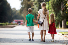 Back View Of Happy Family, Young Blond Long-haired Woman Walking Holding Hands With Two Children, Small Daughter And Son Along Sunny Park Alley On Green Trees Bokeh Background On Warm Summer Day.