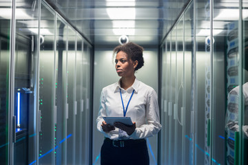 African American woman using tablet while walking in corridor of data center and checking hardware on server racks