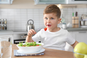 Poster - Adorable little boy eating vegetable salad at table in kitchen