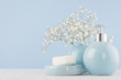 Elegant acessories for dressing table - soft pastel blue ceramic bowls, white flowers, products for skin and body care on white wood board and blue wall, closeup.