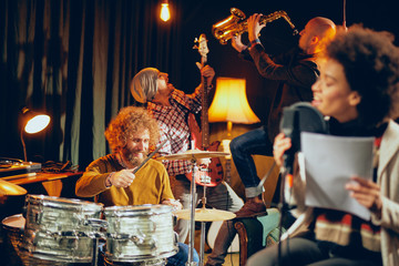 Wall Mural - Mixed race woman singing. In background band playing instruments. Home studio interior. Selective focus on band.