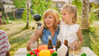 © Gorodenkoff - Granddaughter is Sitting on her Grandmother's Feet. Big Family Garden Party Celebration, Gathered Together at the Table, Eating, Joking and Having Fun.