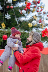 Canvas Print - A young, stylish father wearing a red jacket and hat-ears hugs, kisses and throws up his baby. New Year's photo near a Christmas tree in a city park.