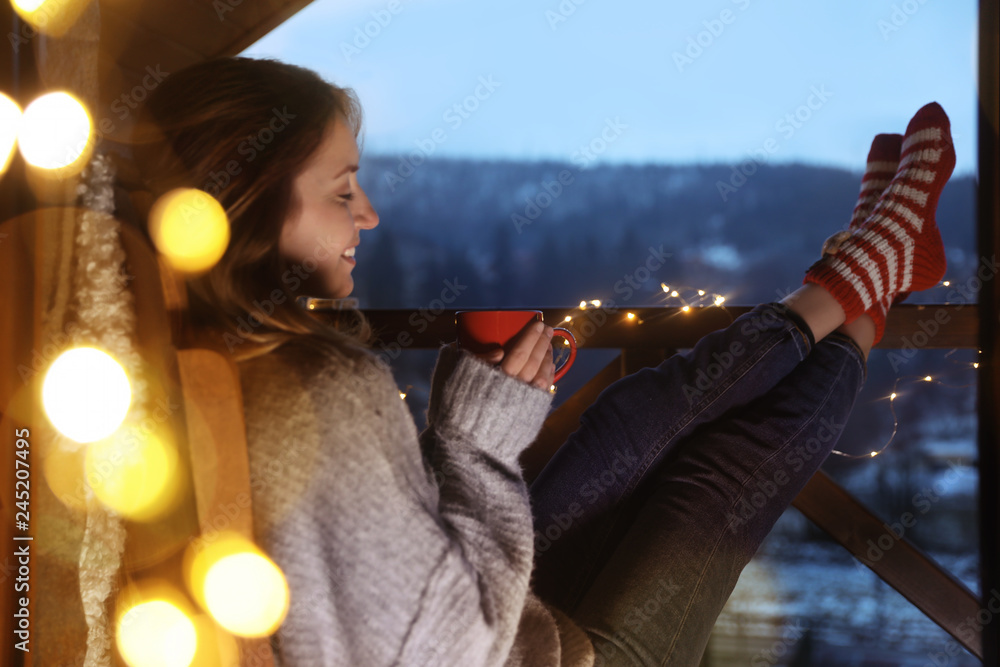 Woman with cup of hot beverage and Christmas lights resting on balcony. Winter evening - obrazy, fototapety, plakaty 