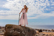 Young woman in long dress posing on rocks  on coast of sea, in hands holds a Viking sword