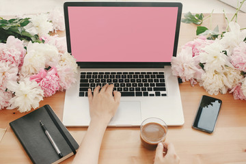 Hands working on stylish laptop with empty screen, coffee, phone, black notebook and peonies on rustic wooden table. Freelance concept. Space for text.  Women's Day. Mothers day