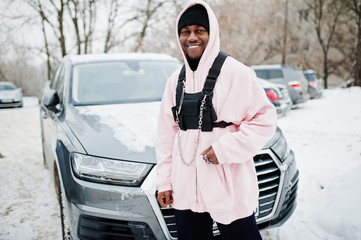 Wall Mural - Stylish urban style african american man in pink hoodie posed against suv car at winter. Afro rapper guy.