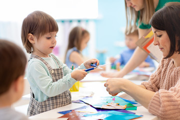 Child with scissors in hands cutting paper with teachers in class room. Group of children doing project in kindergarten. Kids play into team.