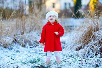 Outdoor winter portrait of little cute toddler girl in red coat and white fashion hat barret. Healthy happy baby child walking in the park on cold day with snow and snowfall. Stylish clothes for kids.