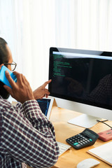 Wall Mural - Modern it developer sitting in front of computer screen with code and talking by smartphone at workplace