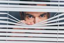 Suspicious Young Man Looking At Camera Through Blinds, Mistrust Concept
