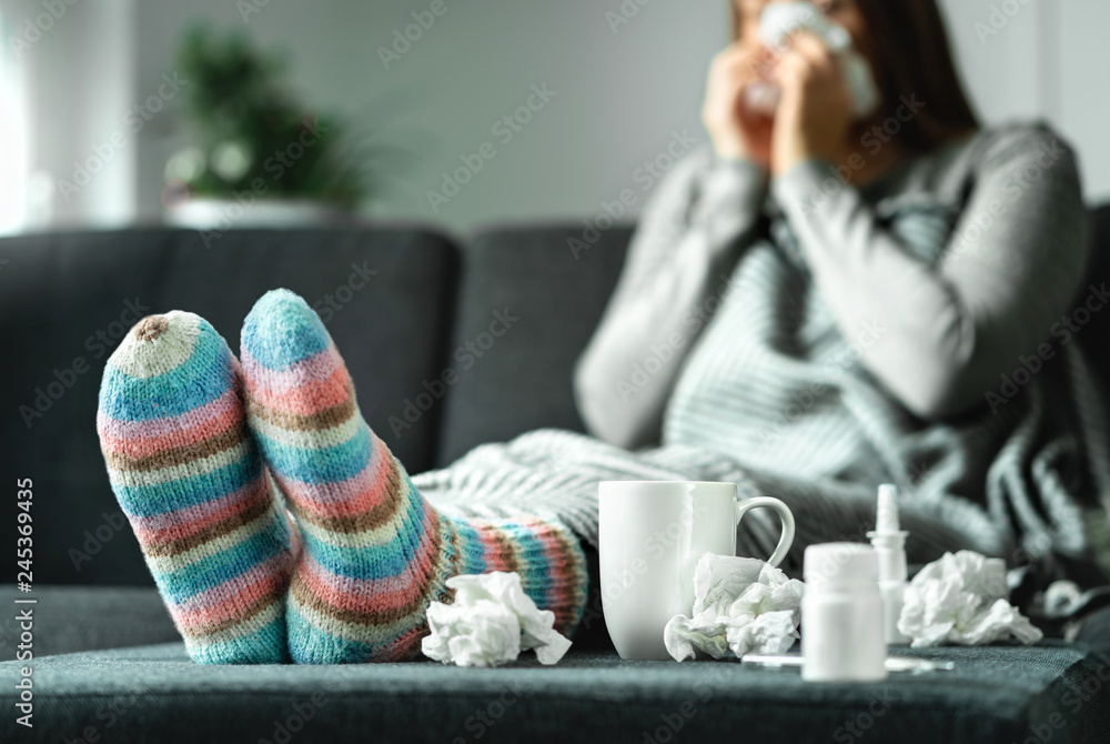 Sick woman with flu, cold, fever and cough sitting on couch at home. Ill person blowing nose and sneezing with tissue and handkerchief. Woolen socks and medicine. Infection in winter. Resting on sofa. - obrazy, fototapety, plakaty 