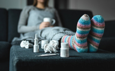 Sick woman resting on couch holding hot cup of tea. Ill person with flu, cold, fever or virus sitting on sofa at home in winter. Medicine, thermometer, nasal spray and woolen socks. Medical concept.