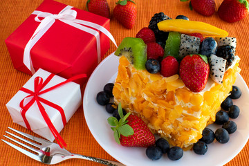 Beautiful, delicious, fruit cake in white plate, red and white gift boxes, fork on orange tablecloth.