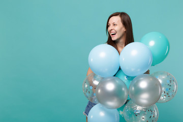 Joyful laughing young woman in denim clothes looking aside, celebrating and holding colorful air balloons isolated on blue turquoise wall background. Birthday holiday party, people emotions concept.