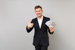 © ViDi Studio - Attractive young business man in suit showing thumb up, holding lots bunch of dollars banknotes, cash money isolated on grey background. Achievement career wealth business concept. Mock up copy space.