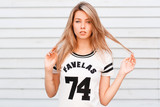 Cute young girl with beautiful blue eyes with sexy lips with natural make-up in a stylish white t-shirt posing on the street near the white wooden wall on a summer day. American stylish girl.