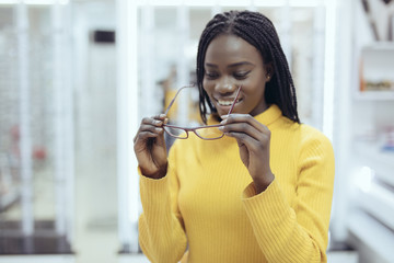 Young african woman choosing eyeglasses frames in Optical Store. Cute girl deciding for glasses of different shapes and colors