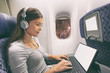 © Maridav - Plane passenger business woman professional working in airplane cabin during flight with in-flight wifi typing writing on tablet computer listening to music with headphones. Young Asian traveler.