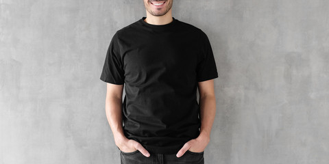 Wall Mural - Attractive young man with poses in blank black cotton t-shirt, standing against gray textured wall