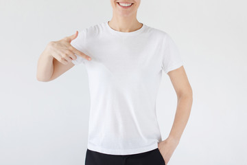 Wall Mural - Woman pointing at her blank white t-shirt with index finger, copy space for your advertising, isolated on gray background. No face photo