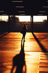 Wall Mural - Back view silhouette of a young girl in warm morning sunrise light and long shadows on the ground on a cold winter day in a park house steel architecture street view. Köln, Cologne in Germany