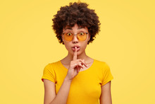 Stupefied Secret Dark Skinned Woman Makes Gesture Quietly, Asks Remain Silent, Gossips About Something, Looks Mysteriously As Tells Secret, Wears Bright Yellow T Shirt, Stands Indoor. Shh, Dont Speak