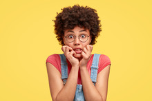 Headshot Of Frustrated Mixed Race Young Woman Has Crisp Hair, Bites Finger Nails With Nervous Expression, Stares With Eyes Full Of Fear, Wears Casual Clothes, Stands Against Yellow Studio Wall