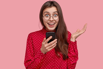 Headshot of cheerful European girl holds cellular, stays in touch, sends message, happy to recieve feedback, spreads palm, poses against pink background, explores social network joins online community