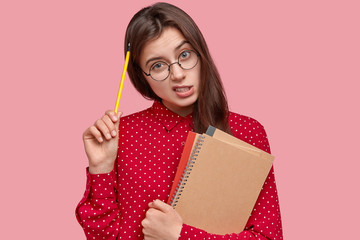 Wall Mural - Daily schedule and checklists of tasks. Puzzled young woman scratches head with pencil, thinks what is done, writes notes in notepad, has thoughtful expression, isolated over pink background