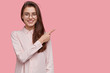 © wayhome.studio  - Delighted female has cheerful expression, points with fore finger at blank space, dressed in elegant formal shirt, stands against pink background with copy space for your promotional content.