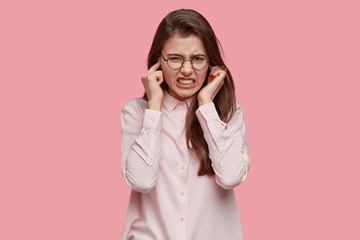 Wall Mural - Frustrated young lady plugs ears, clenches teeth from annoyance, ignores annoying sound, wears transparent spectacles, hears noise coming from neighbours, isolated over pink background. Decibel