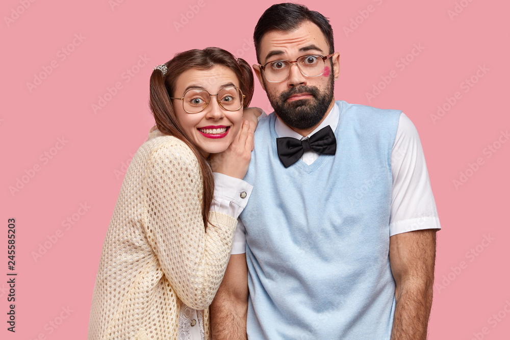 Portrait of two clumsy female and male wonks stand closely, wear spectacles, pose for making common photo, express happiness and surprisement, isolated over pink background. Geek couple indoor
