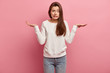 Studio shot of puzzled female shrugs shoulders, has uncertain facial expression, can not understand what is better, wears casual white sweatshirt and jeans, models indoor over pink studio wall