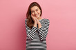 © wayhome.studio  - Studio shot of attractive woman with healthy skin, pleasant appearance, keeps hands together near cheek, has dark hair, smiles gently, wears casual striped clothes, isolated over pink background