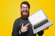 © Vulp - Happy bearded man in glasses holding and pointintg at laptop while looking at camera