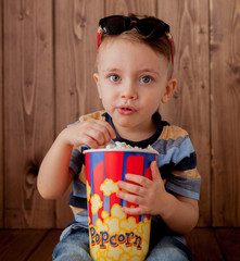 Little cute kid baby boy 2-3 years old , 3d cinema glasses holding bucket for popcorn, eating fast food on wooden background. Kids childhood lifestyle concept. Copy space