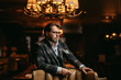 Portrait of wealthy prosperious handsome businessman, dressed in tailored three-piece suit sitting in leather arm-chai at elite gentlemen club with lighted multi-lamp chandelier behind.