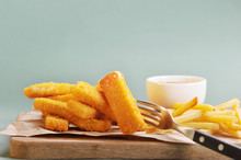 Fish Sticks And French Fries On A Light Wooden Board. Sauce Is Used As A Supplement. Fish Stick Pierced With A Fork. Light Background. Close-up. 