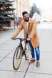 © F8  \ Suport Ukraine - Happy young indian man dressed in shirt and coat standing with a fixed gear bicycle on city street