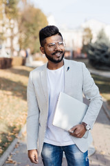 Wall Mural - Handsome young indian hipster man holding his laptop while standing on the city street