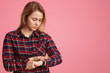 Serious European woman hurries on meeting, looks at watch, checks time, dressed in checkered shirt, poses against pink background with blank space for your promotional content. Its time to go