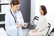 © rogerphoto - Doctor woman at work. Portrait of female physician filling up medical form while standing near reception desk at clinic or emergency hospital. Patient woman sitting at the background. Medicine concept
